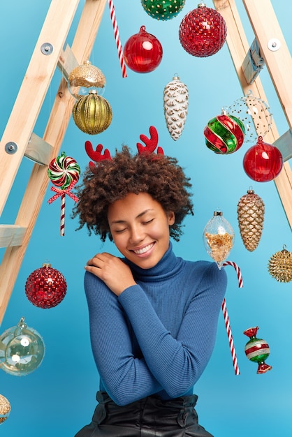 Foto vertical de uma mulher afro-americana satisfeita desfrutando de uma atmosfera mágica festiva em casa usando poses confortáveis de gola alta