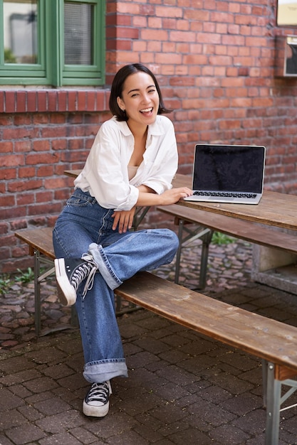 Foto grátis foto vertical de uma mulher asiática bonita com laptop sentado em um café ao ar livre tomando café e