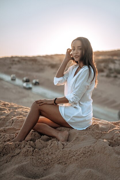 Foto vertical de uma mulher branca em um vestido branco posando na praia