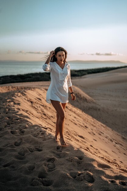 Foto vertical de uma mulher branca em um vestido branco posando na praia