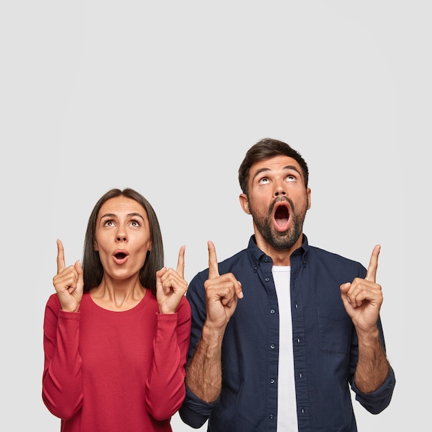 Foto vertical de uma mulher caucasiana surpresa e um homem apontando com os dois dedos indicadores para cima, mostra espaço livre para promoção