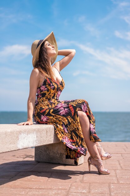 Foto vertical de uma mulher com um vestido de verão floral e um chapéu sentada à beira-mar, capturada na Espanha