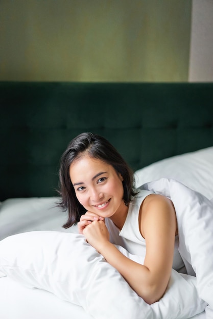 Foto grátis foto vertical de uma mulher coreana sorridente deitada no travesseiro em sua cama, passando a manhã no quarto acordando