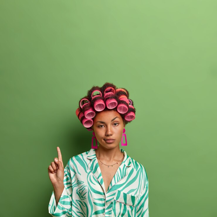 Foto grátis foto vertical de uma mulher de pele escura não impressionada usando bobes de cabelo e indica no espaço da cópia, mostra o espaço em branco contra a parede verde para sua promoção, se preocupa com a aparência, se prepara para o encontro