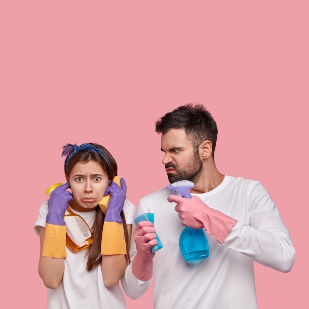 Foto vertical de uma mulher descontente tapa os ouvidos, não quer ouvir o marido zangado que reclama de muito trabalho doméstico, segura sprays, esponjas, faz limpeza de primavera, usa luvas de proteção, roupas brancas