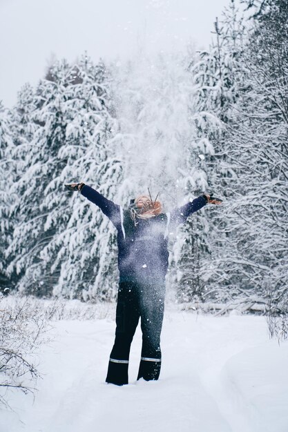 Foto vertical de uma mulher jogando neve para o alto em uma floresta de inverno