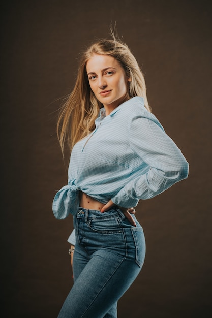 Foto vertical de uma mulher loira atraente em jeans e uma camisa curta posando em um fundo marrom