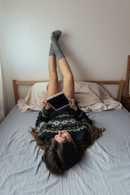 Foto vertical de uma mulher usando seu tablet deitada na cama com as pernas na parede