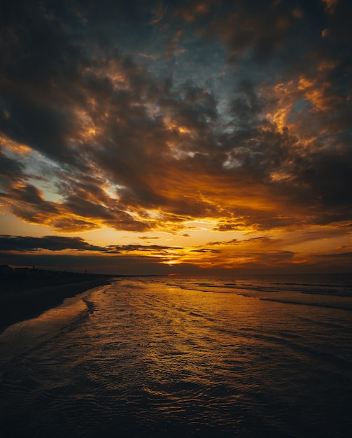 Foto vertical de uma praia cercada pelas ondas do mar sob um céu nublado durante um belo pôr do sol