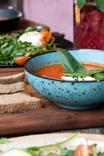 Foto vertical de uma tigela de sopa de tomate e um prato de salada e pão em uma placa de madeira