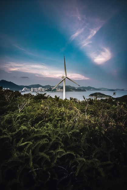 Foto vertical de uma turbina eólica em um campo com o mar ao fundo