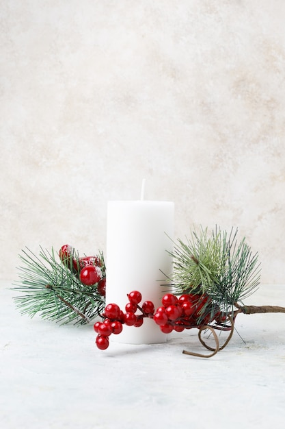 Foto vertical de uma vela branca cercada por azevinhos de Natal e folhas em uma superfície de mármore branco