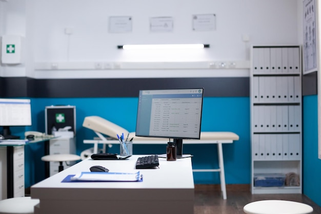 Fotografia ampla de uma sala médica vazia, moderna e limpa no interior do hospital. Computador na mesa com lista de dados de consultas e equipamentos de escritório clínico no espaço médico do médico