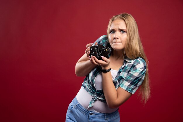 Fotografia de jovem loira segurando uma câmera profissional e não sabe como usá-la.