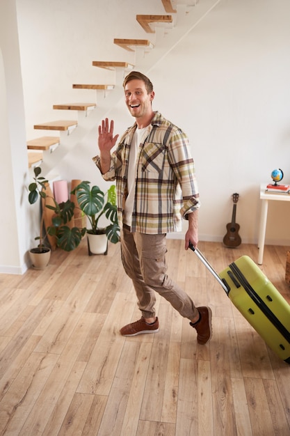 Foto grátis fotografia vertical de um jovem feliz com uma mala a verificar no apartamento alugado a agitar a mão para