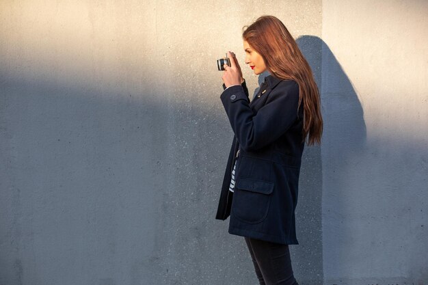 Fotógrafo feminino sorridente na jaqueta em frente à parede pronta para fazer uma nova foto. Adorável jovem morena com roupa da moda posando no fundo da parede de concreto com câmera