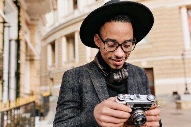 Fotógrafo masculino sorridente em fones de ouvido em pé na rua da cidade. foto ao ar livre de alegre jovem africano com jaqueta quadriculada, olhando para a câmera.