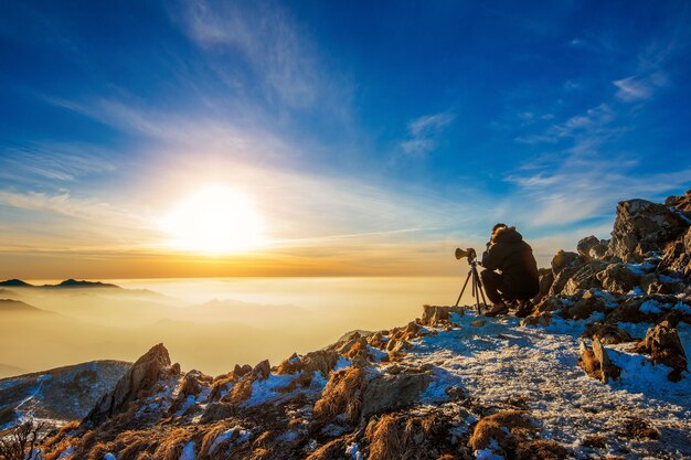 Fotógrafo profissional tira fotos com a câmera no tripé no pico rochoso ao pôr do sol