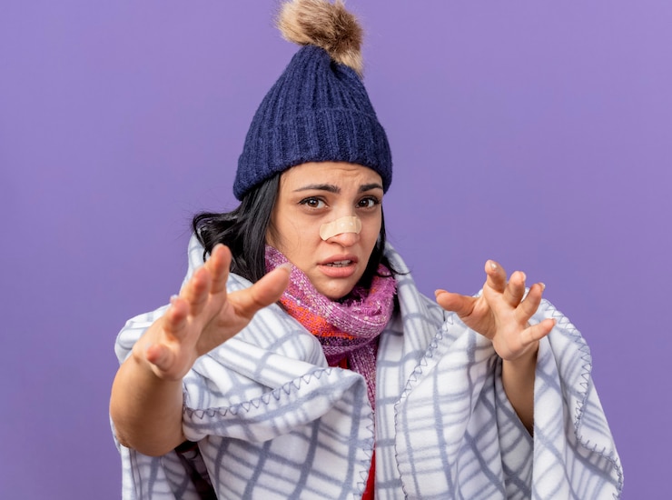 Foto grátis fraca jovem caucasiana doente com chapéu de inverno e lenço embrulhado em xadrez olhando para a câmera com gesso médico no nariz, estendendo as mãos em direção à câmera