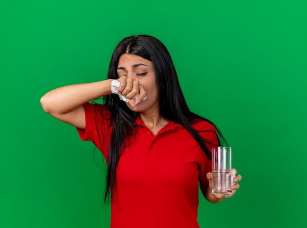 Fraca jovem caucasiana doente segurando um pacote de comprimidos, copo de água e guardanapo se preparando para espirrar, mantendo a mão no nariz com os olhos fechados, isolado na parede verde com espaço de cópia