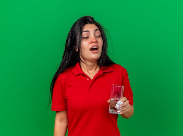 Fraca jovem caucasiana doente segurando um pacote de comprimidos, um copo de água se preparando para espirrar com os olhos fechados, isolado em um fundo verde com espaço de cópia