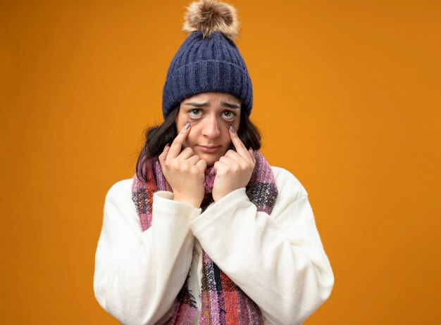 Fraca jovem caucasiana doente usando um manto de inverno, chapéu e cachecol, olhando para a câmera, puxando para baixo as pálpebras dos olhos, isoladas em um fundo laranja com espaço de cópia