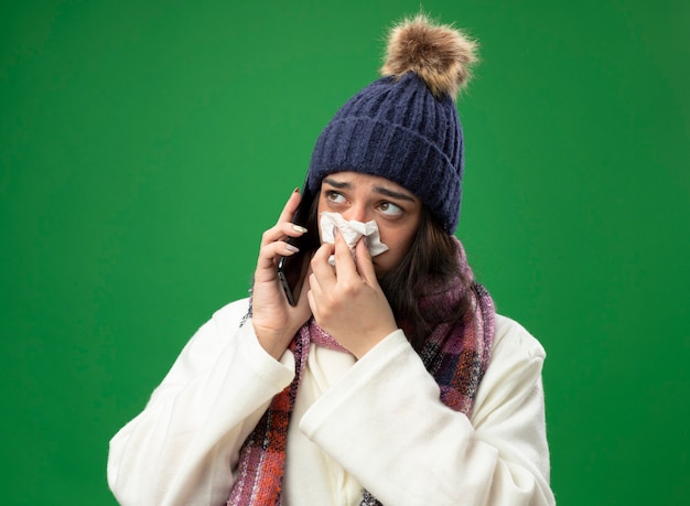 Fraca jovem caucasiana doente usando um manto de inverno, chapéu e lenço falando no telefone, limpando o nariz com guardanapo, olhando para o lado isolado no fundo verde
