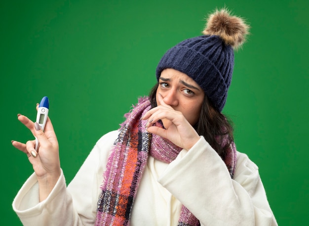 Fraca jovem caucasiana doente usando um manto de inverno, chapéu e lenço segurando um termômetro, limpando o nariz isolado na parede verde
