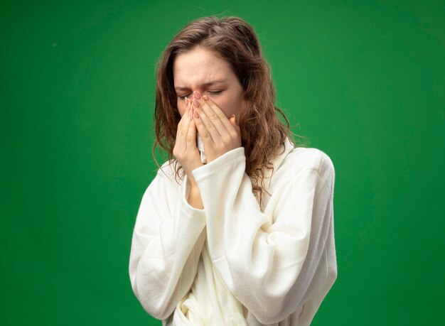 Fraca jovem doente com os olhos fechados, usando um manto branco, limpando o nariz com um guardanapo isolado no verde