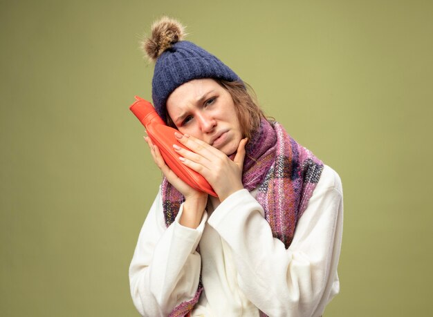 Fraca jovem doente vestindo túnica branca e chapéu de inverno com lenço segurando uma bolsa de água quente na bochecha isolada em verde oliva