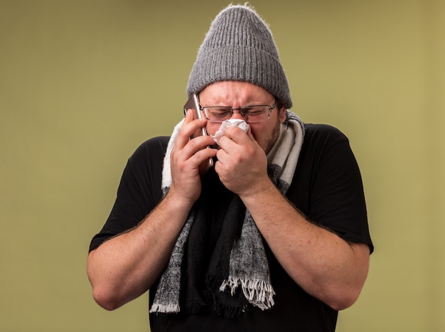 Fraco homem doente de meia-idade usando chapéu de inverno e lenço fala no telefone limpando o nariz com guardanapo isolado na parede verde oliva