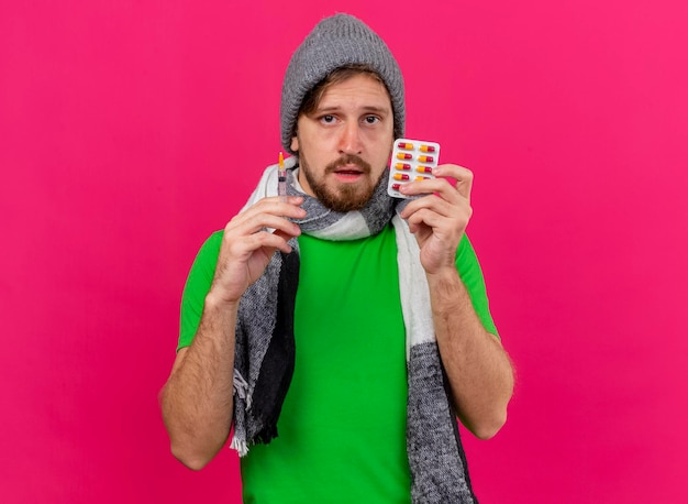 Fraco, jovem, bonito, eslavo, homem, usando, inverno, chapéu e, lenço, segurando, seringa e, pacote, cápsulas, olhando para frente