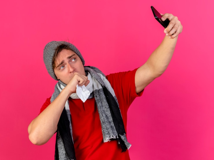 Foto grátis fraco jovem bonito loira doente com chapéu de inverno e lenço segurando o guardanapo, mantendo a mão na boca, tirando uma selfie isolada no fundo carmesim