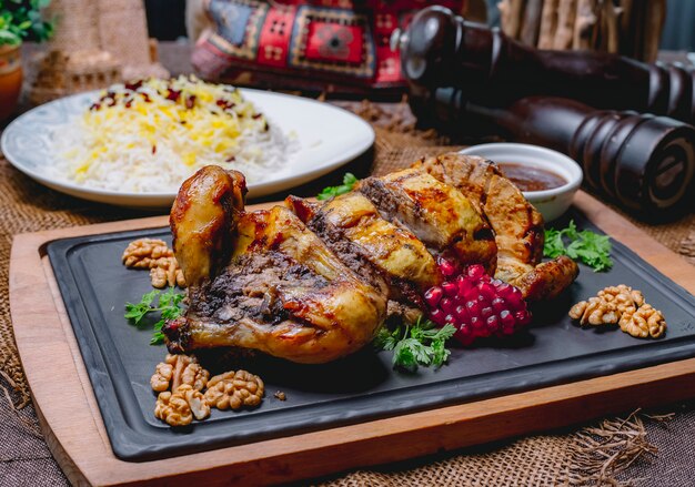 Frango recheado assado, decorado com romã e nozes em uma placa preta e arroz em um prato branco sobre uma mesa de madeira