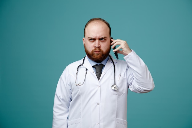 Franzindo a testa jovem médico masculino vestindo jaleco e estetoscópio em volta do pescoço, olhando para o lado falando no telefone isolado em fundo azul