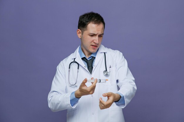 Franzindo a testa jovem médico masculino vestindo roupão médico e estetoscópio no pescoço, mantendo a mão no ar segurando a seringa com agulha olhando para o braço isolado no fundo roxo