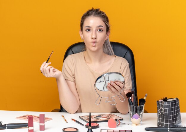 Franzindo os lábios, bela jovem se senta à mesa com ferramentas de maquiagem segurando um pincel de maquiagem com espelho isolado em um fundo laranja