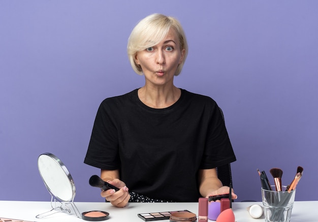 Franzindo os lábios, uma jovem linda se senta à mesa com ferramentas de maquiagem segurando um blush em pó isolado sobre um fundo azul