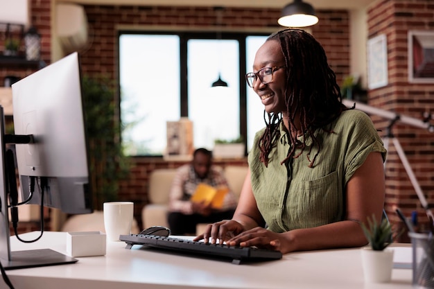 Freelancer americano africano trabalhando digitação remota e olhando para a tela do computador enquanto o namorado está relaxando. Programador sorridente usando pc para conversar com amigos enquanto colega de quarto está lendo um livro.