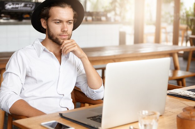 Freelancer barbudo conectando-se à rede sem fio via laptop. Homem pensativo, trabalhando no notebook enquanto está sentado à mesa de madeira no interior da cafeteria moderna. Livro de leitura do aluno em café