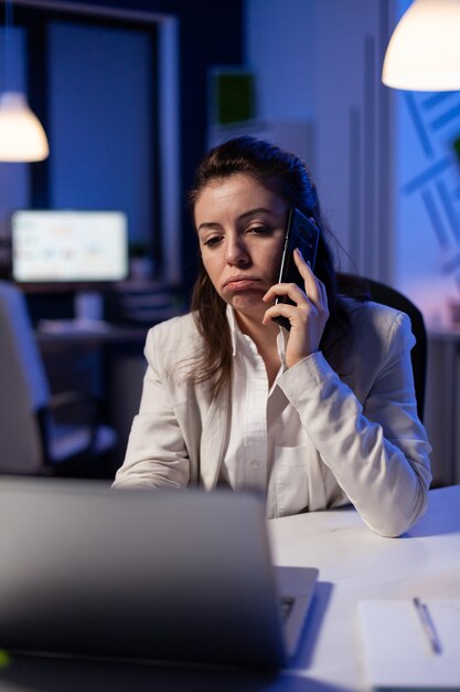 Freelancer corporativo cansado conversando no telefone enquanto trabalhava exausto para estatísticas econômicas em um escritório de negócios tarde da noite. Funcionário focado em uma rede de tecnologia moderna sem fio