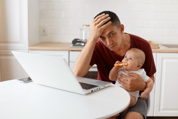 Freelancer de homem morena atraente exausto, vestindo camiseta marrom estilo casual, trabalhando e cuidando de sua filha bebê, mantendo a mão em sua testa.