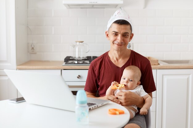 Freelancer engraçado positivo morena atraente vestindo camiseta marrom estilo casual e fralda na cabeça, trabalhando e cuidando de sua filha bebê.