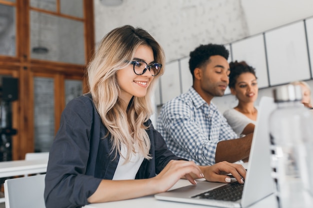 Freelancer feminino encaracolado lindo com manicure bonita usando laptop e sorrindo. retrato interior da secretária loira sentada ao lado de um colega de trabalho africano de camisa azul.