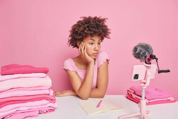 Freelancer feminino encaracolado sério e atencioso assiste ao vídeo como lavar a roupa em casa, senta-se à mesa com a roupa dobrada isolada sobre o rosa