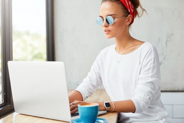 Freelancer feminino jovem trabalha remotamente no computador portátil no café, informações de teclados, bebe café. Mulher conversa com amigos em redes sociais, conectada à internet sem fio em restaurante