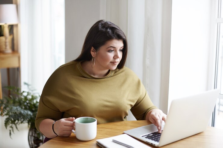 Foto grátis freelancer jovem atraente gordinho vestindo um suéter elegante e brincos redondos, trabalhando na frente de um laptop aberto, sentado no aconchegante interior do escritório em casa, bebendo café, navegando em sites