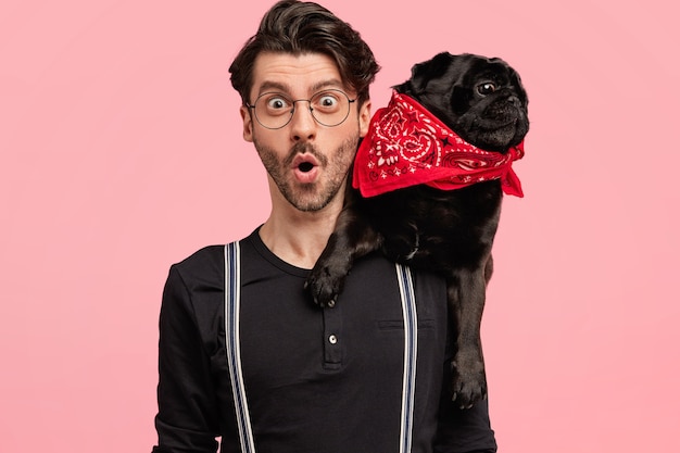 Foto grátis freelancer jovem chocado com aparência específica passa o tempo de lazer na companhia de cachorro, tem expressão de surpresa, olhares fixos e poses contra a parede rosa. conceito de pessoas e animais de estimação