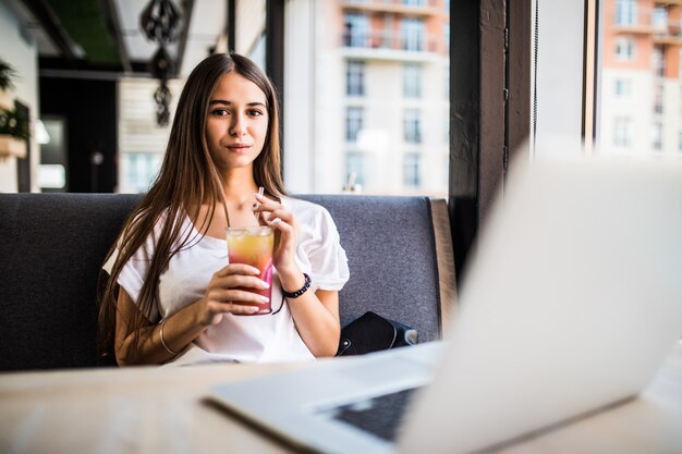 Freelancer linda mulher alegre de bom humor, usando o computador portátil, sentado em cafés, bebendo Mojitos.