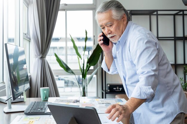 Freelancer masculino asiático sênior idoso em roupas casuais digitando no teclado do laptop enquanto fala no smartphone em pé na mesa e ocupado trabalhando no escritório em casa